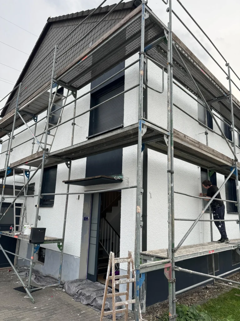Eingerüstetes Einfamilienhaus mit frisch verputzter weißer Fassade, ein Handwerker arbeitet am Gerüst